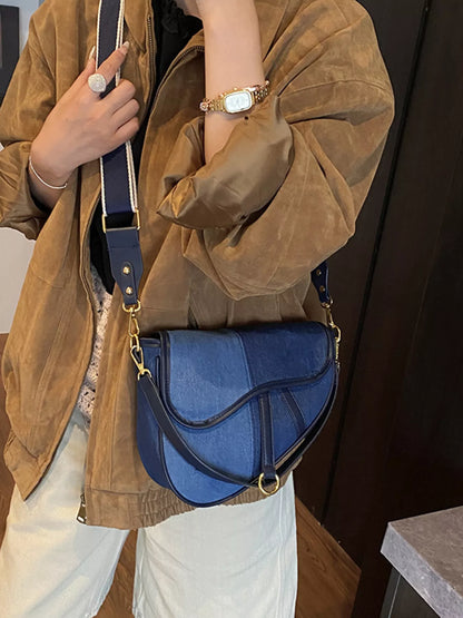 A figure standing in a brown jacket and beige slacks with a crossbody bag composed of two shades of blue denim slung across her body.