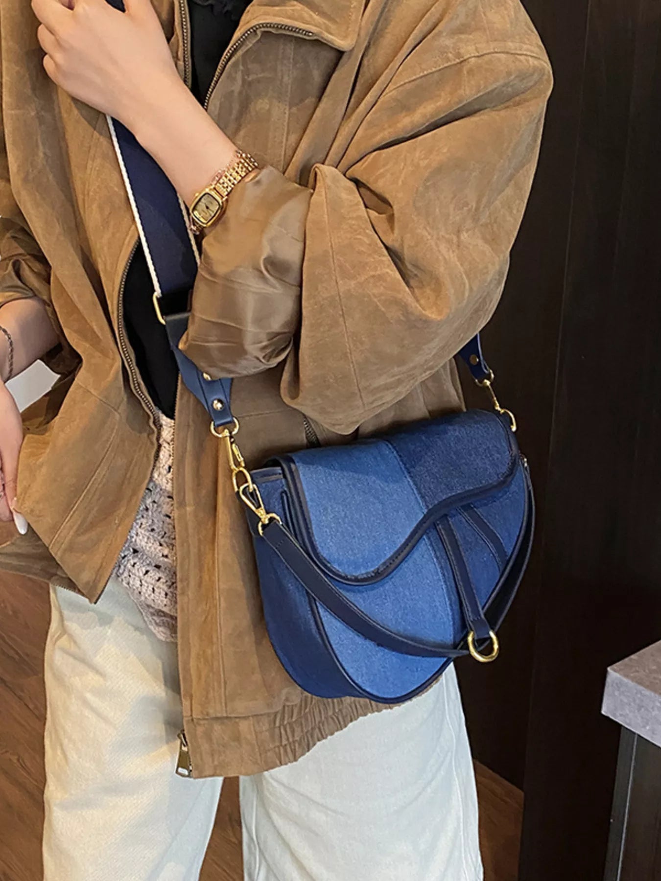 A figure standing in a brown jacket and beige slacks with a crossbody bag composed of two shades of blue denim slung across her body.