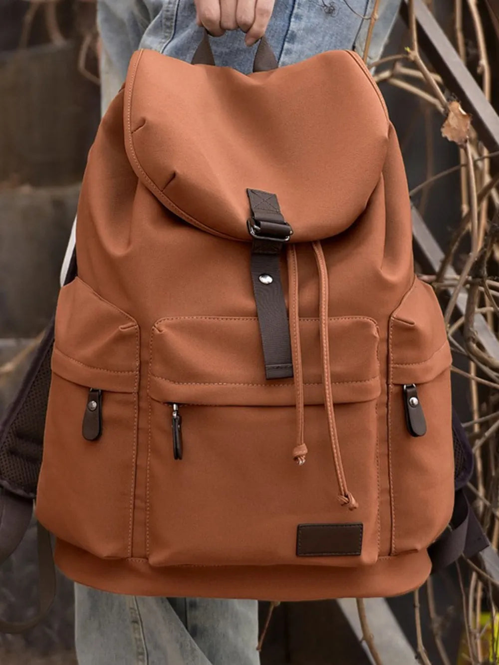 A hand holds a brown backpack with coffee-brown trimming by the top hanging strap. The bag has three lower compartments with zip enclosures - one on either side of the bag and one on the front. The main compartment of the bag has a flap cover.