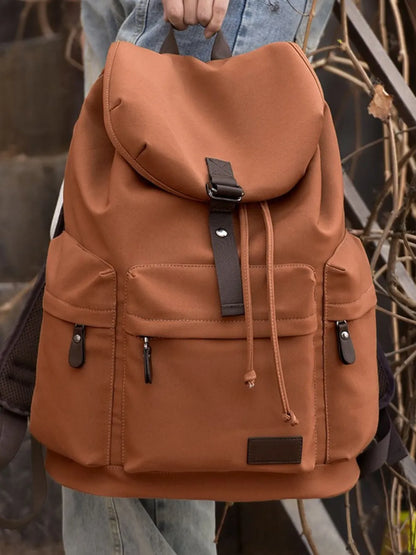 A hand holds a brown backpack with coffee-brown trimming by the top hanging strap. The bag has three lower compartments with zip enclosures - one on either side of the bag and one on the front. The main compartment of the bag has a flap cover.