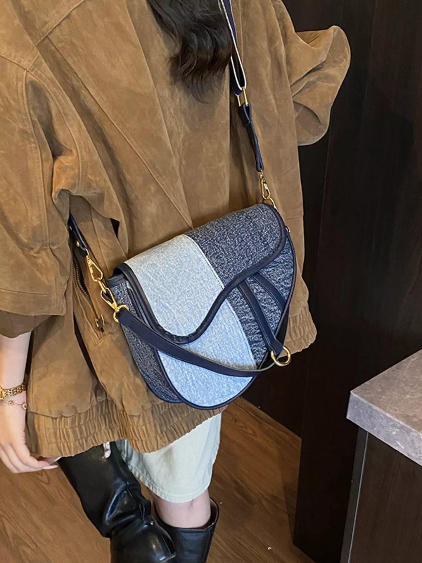 A saddle bag in light blue and dark blue denim on the back of a figure standing in a brown jacket, beige shorts and black boots.