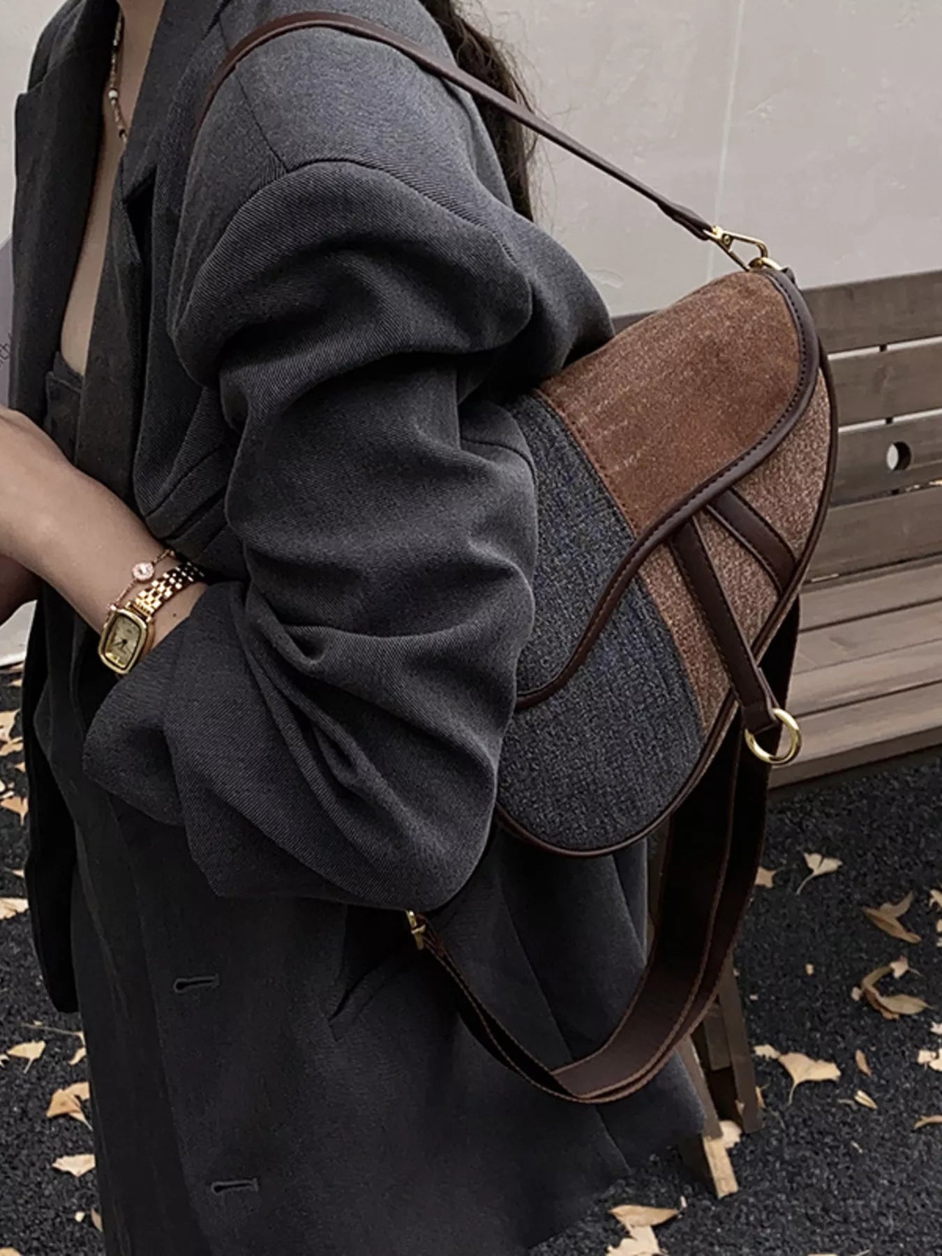 A figure standing outdoors in a dark gray blazer with a dark brown and dark grey two-tone denim bag slung on her left shoulder. 
