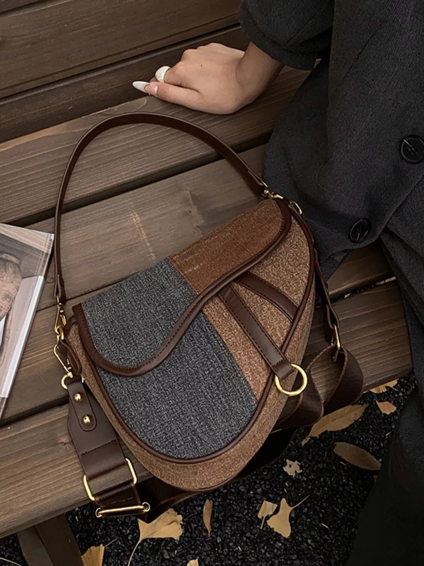 Two-tone brown and dark grey denim saddlebag lying on a brown bench. Also seen in the image are a part of a figure in a gray blazer and a hand.