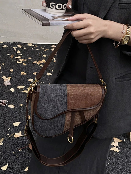 A picture of a figure wearing a dark grey suit holding a book and a cup of beverage in her right hand, and a two-tone brown and grey denim saddlebag in her left hand.   A gold watch and gold bracelet are pictured on her left wrist.